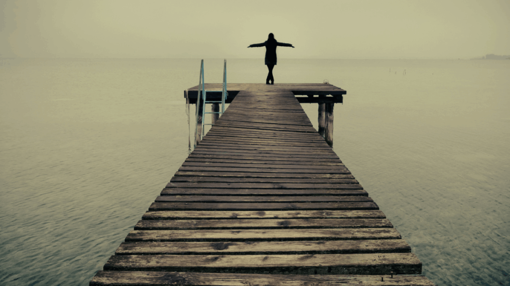 Frau steht am Steg am Wasser und breitet die Arme aus als Symbol für Selbstbestimmung