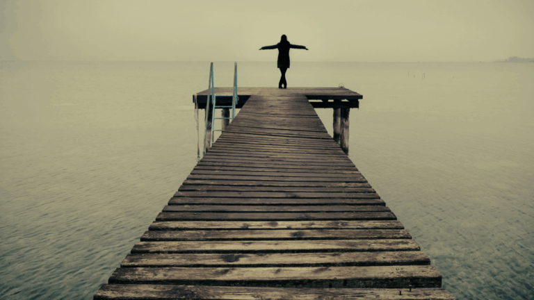 Frau steht am Steg am Wasser und breitet die Arme aus als Symbol für Selbstbestimmung